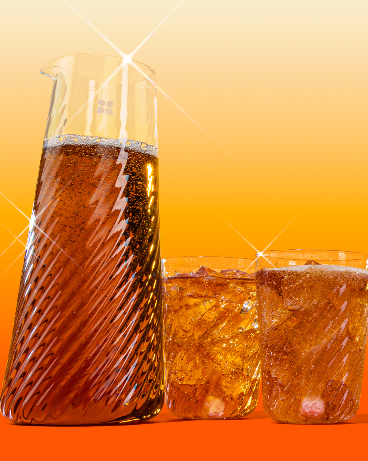 Üvegkancsó és két hullámos pohár sötét szénsavas itallal és jéggel, narancssárga háttér előtt.