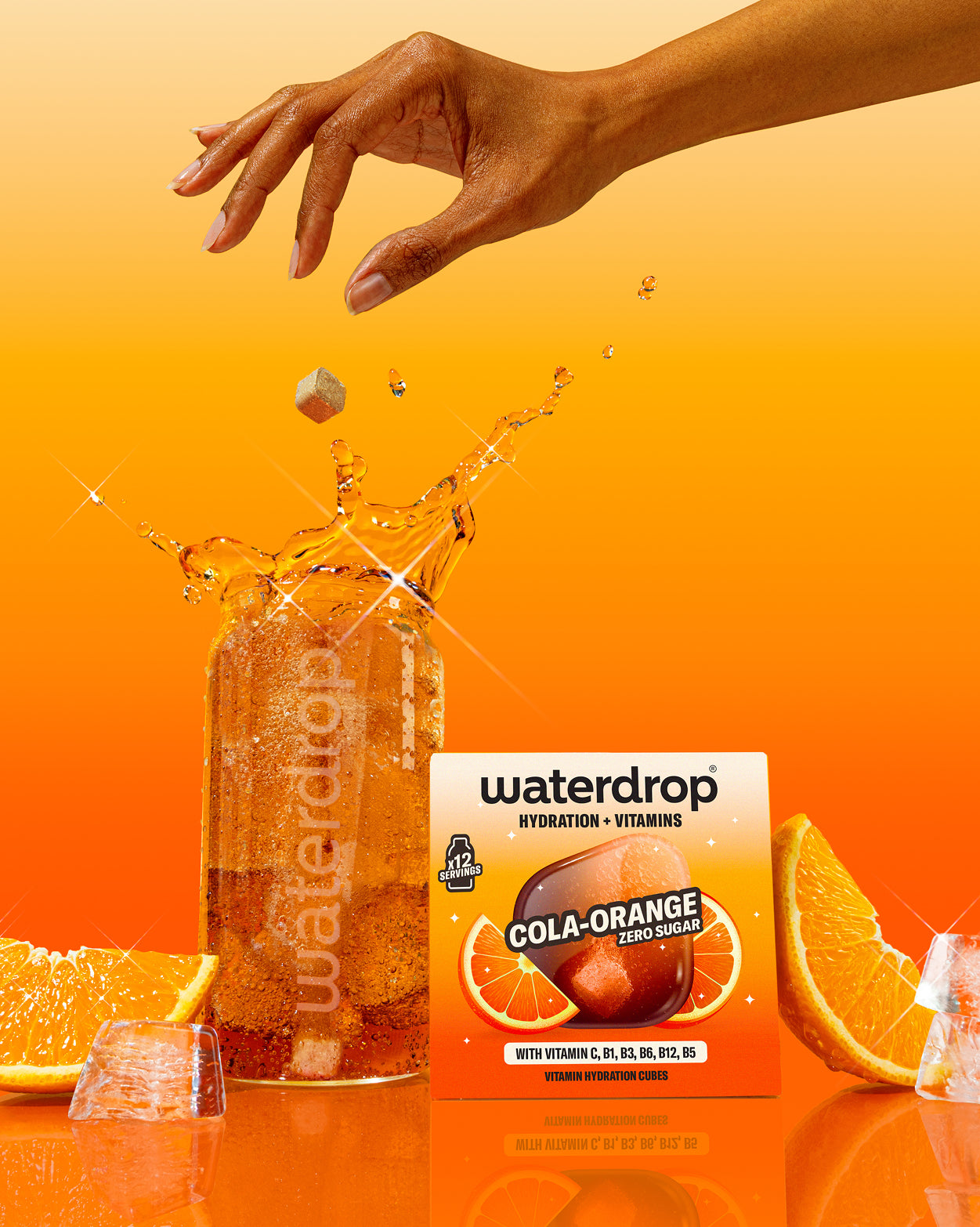 Egy kéz elenged egy Waterdrop Cola Orange kockát, amely egy szénsavas italba esik, a doboz, narancsszeletek és jég mellett, narancssárga háttér előtt.