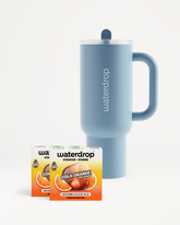 Kék Waterdrop termoszpohár fedéllel és szívószállal, két Cola Orange ízesítésű doboz mellett, fehér háttér előtt.