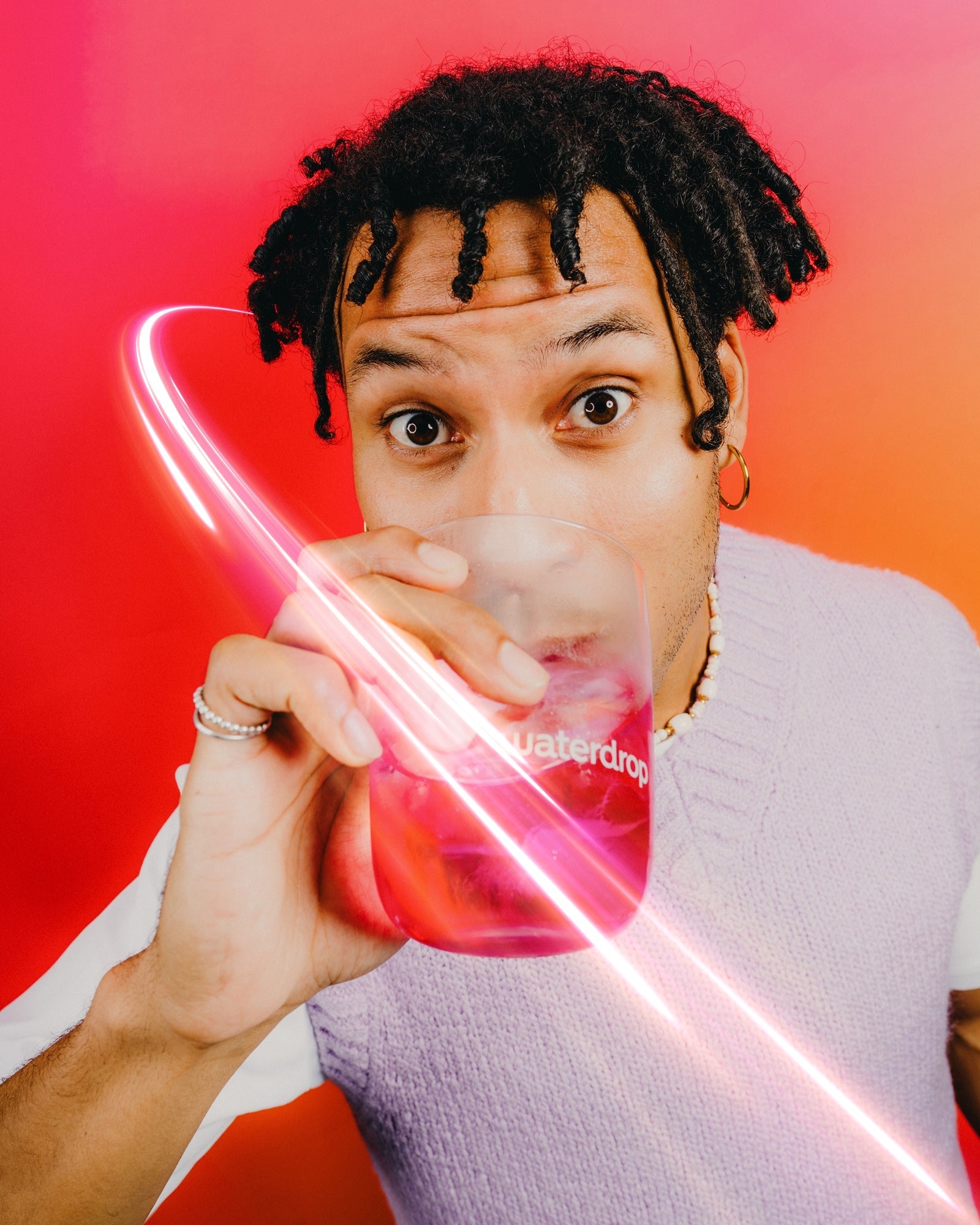Személy, aki egy jégkockás, rózsaszín italos waterdrop pohárból iszik, piros-narancs háttér előtt.