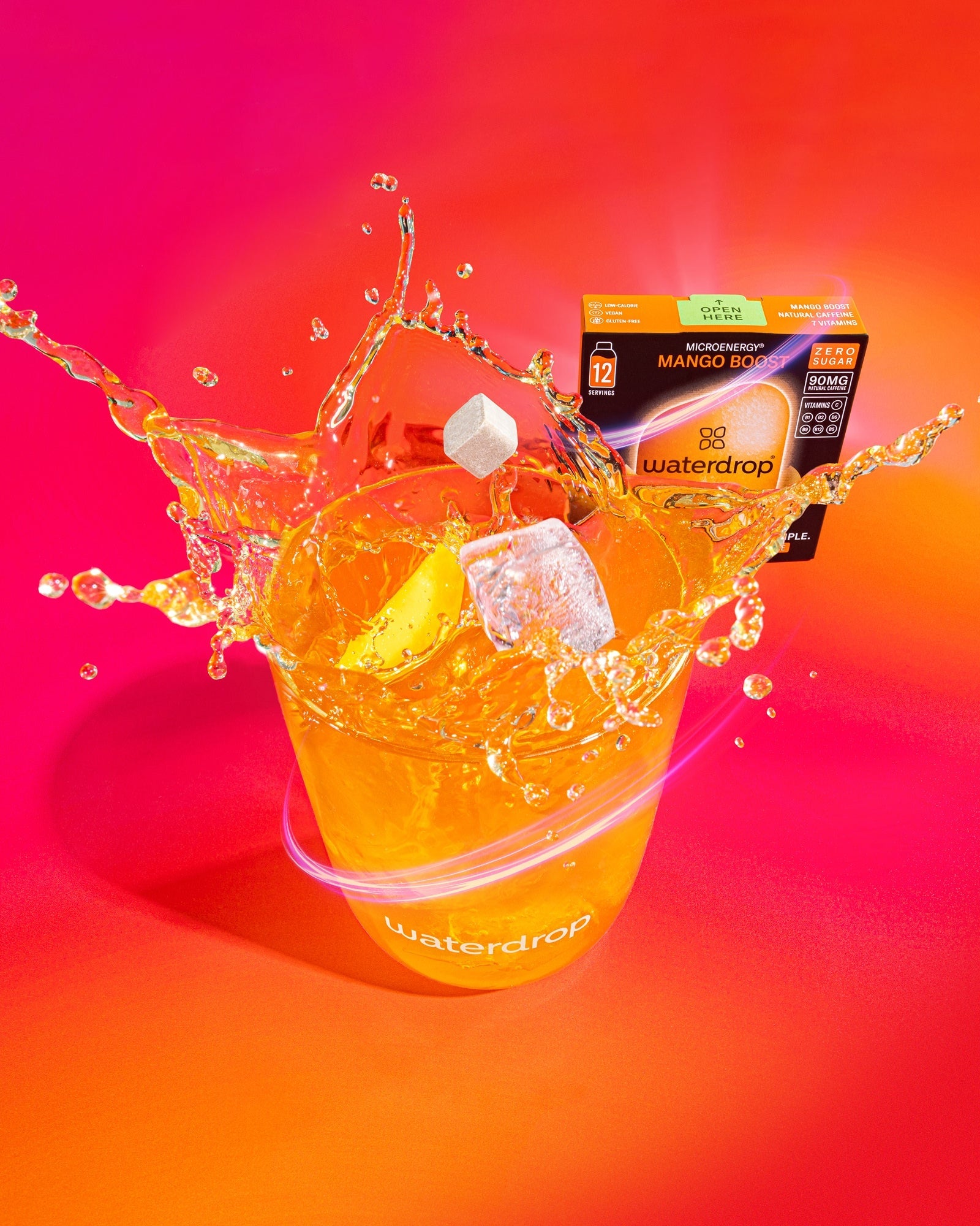 Waterdrop pohár Mango Boost itallal, jégkockákkal és kifröccsenő folyadékkal, mögötte egy cukormentes Microenergy Mango Boost doboz 12 adaggal.