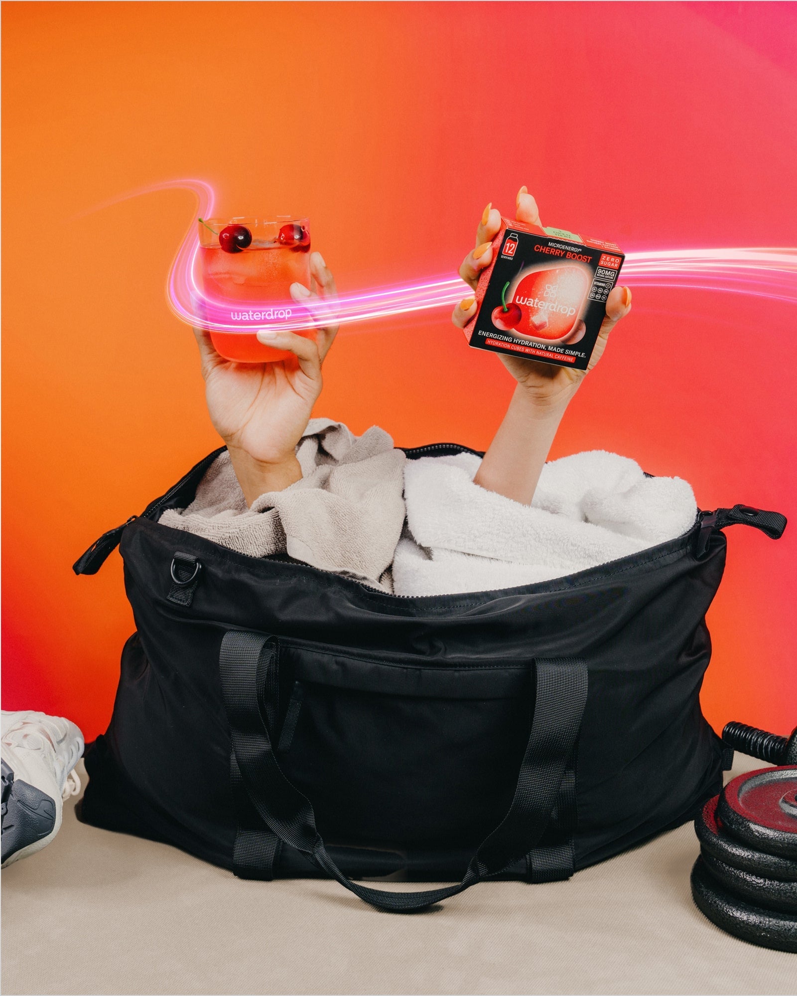 Fekete sporttáskából kilógó kezek, egyikben piros italos waterdrop pohár cseresznyével, a másikban egy cukormentes Microenergy Cherry Boost doboz 12 adaggal, narancssárga háttéren.