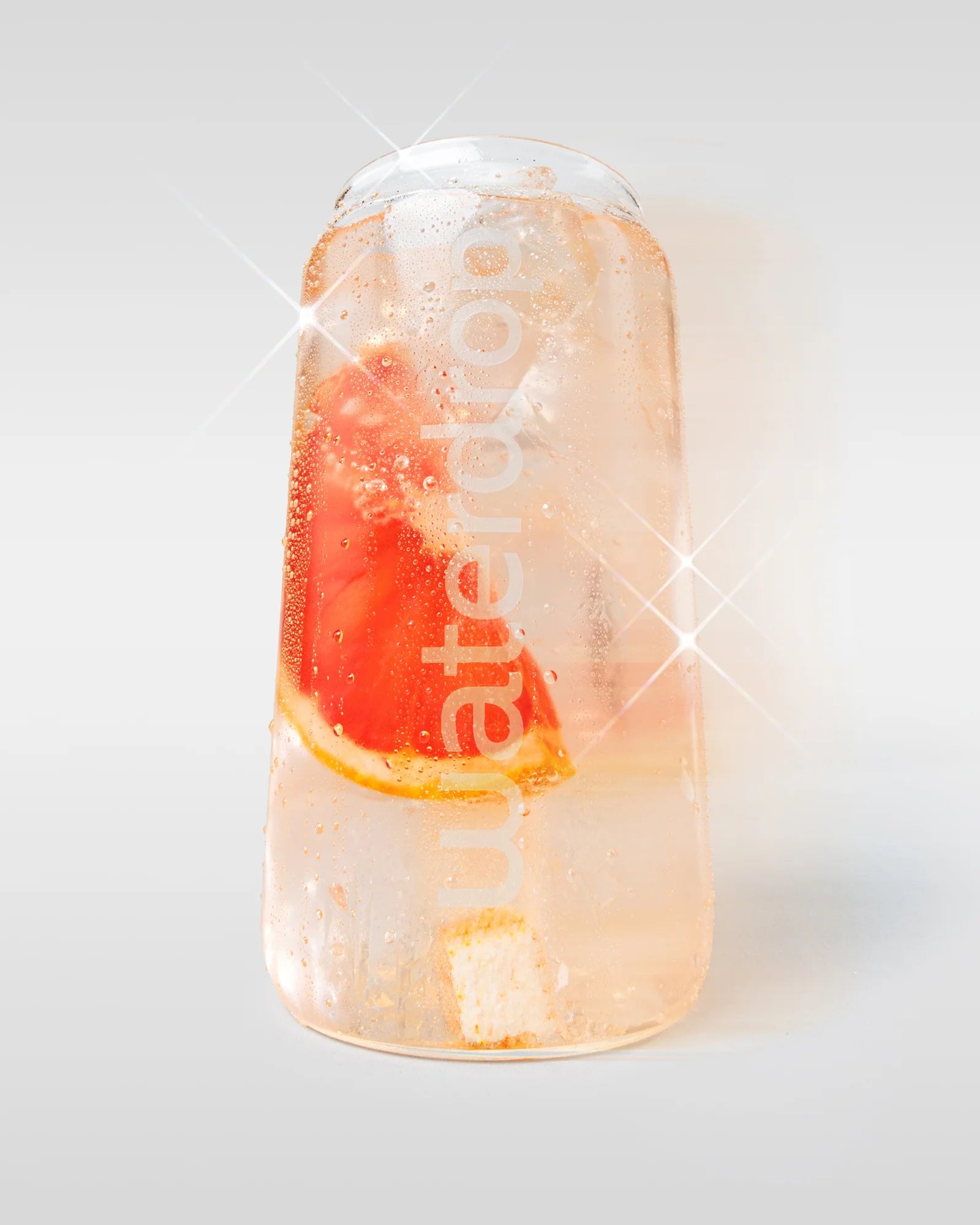 Újrahasználható waterdrop palack világos rózsaszín itallal, jégkockákkal és egy látható grapefruit szelettel, világos háttéren.