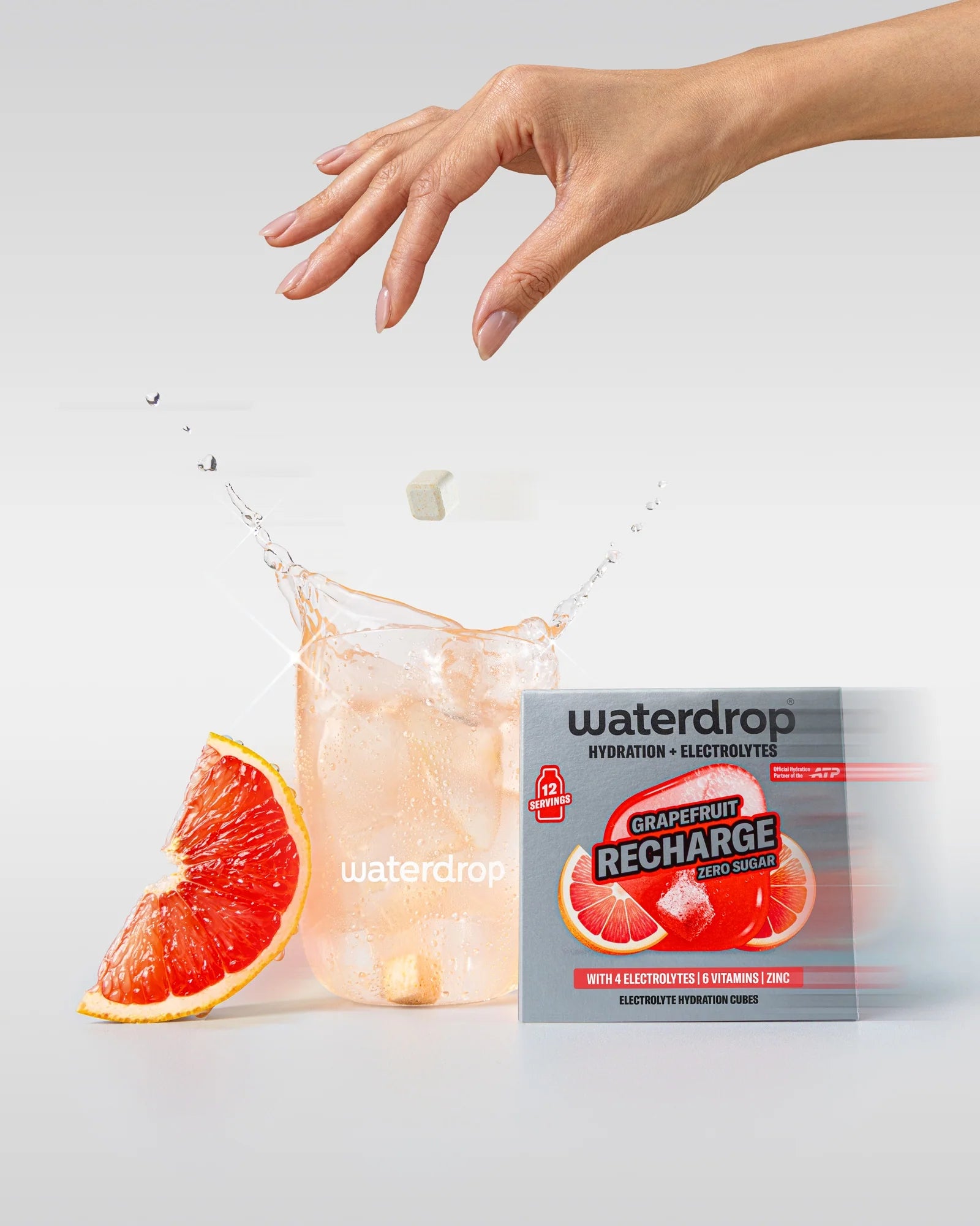 Kéz egy grapefruit italos, jégkockás waterdrop pohár felett, beleeső hidratáló kockával, mellette egy cukormentes Grapefruit Recharge doboz 12 adaggal és egy grapefruit szelet.