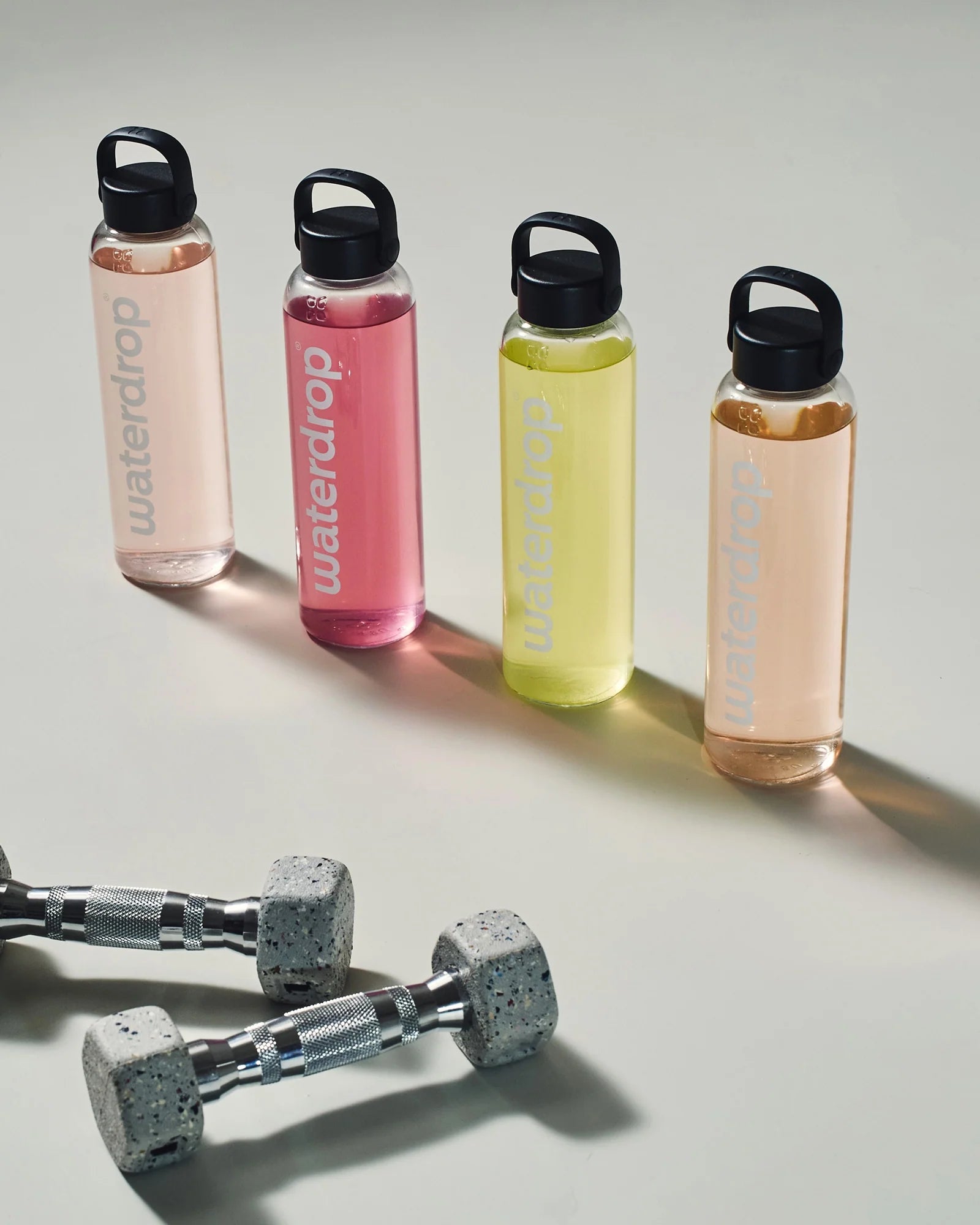 Négy egymás mellett álló újrahasználható waterdrop palack különböző színű italokkal (világos rózsaszín, rózsaszín, sárga és barack), előtérben fém kézisúlyzókkal, világos háttéren.
