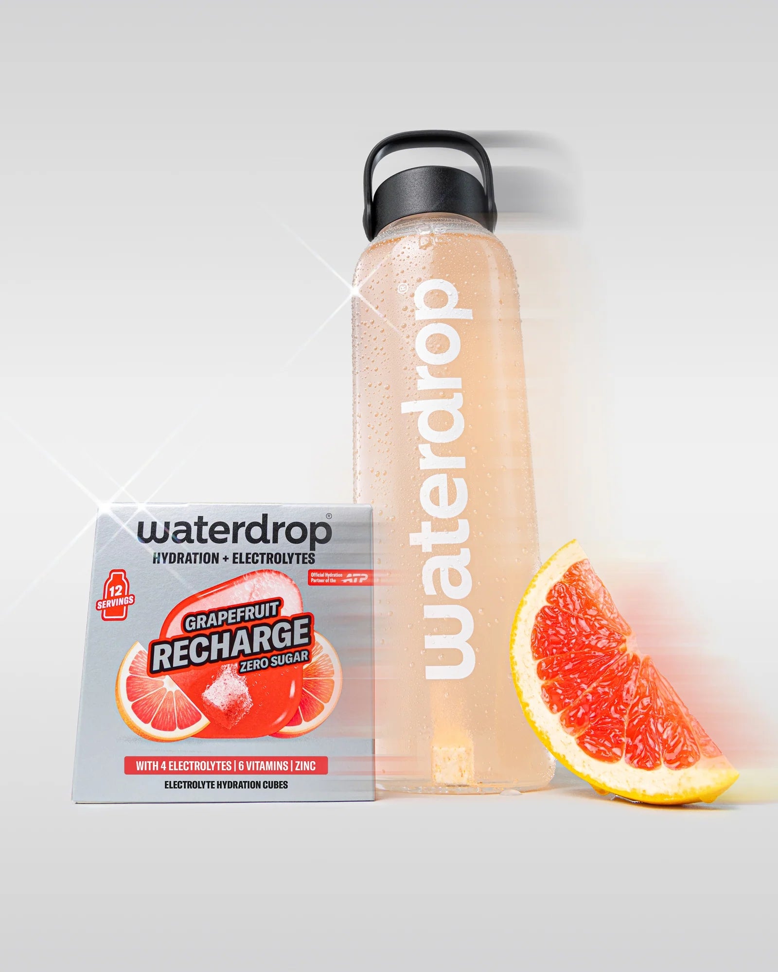 Újrahasználható waterdrop palack rózsaszín grapefruit itallal, mellette egy cukormentes Grapefruit Recharge doboz 12 adaggal és egy grapefruit szelet, fehér háttéren.