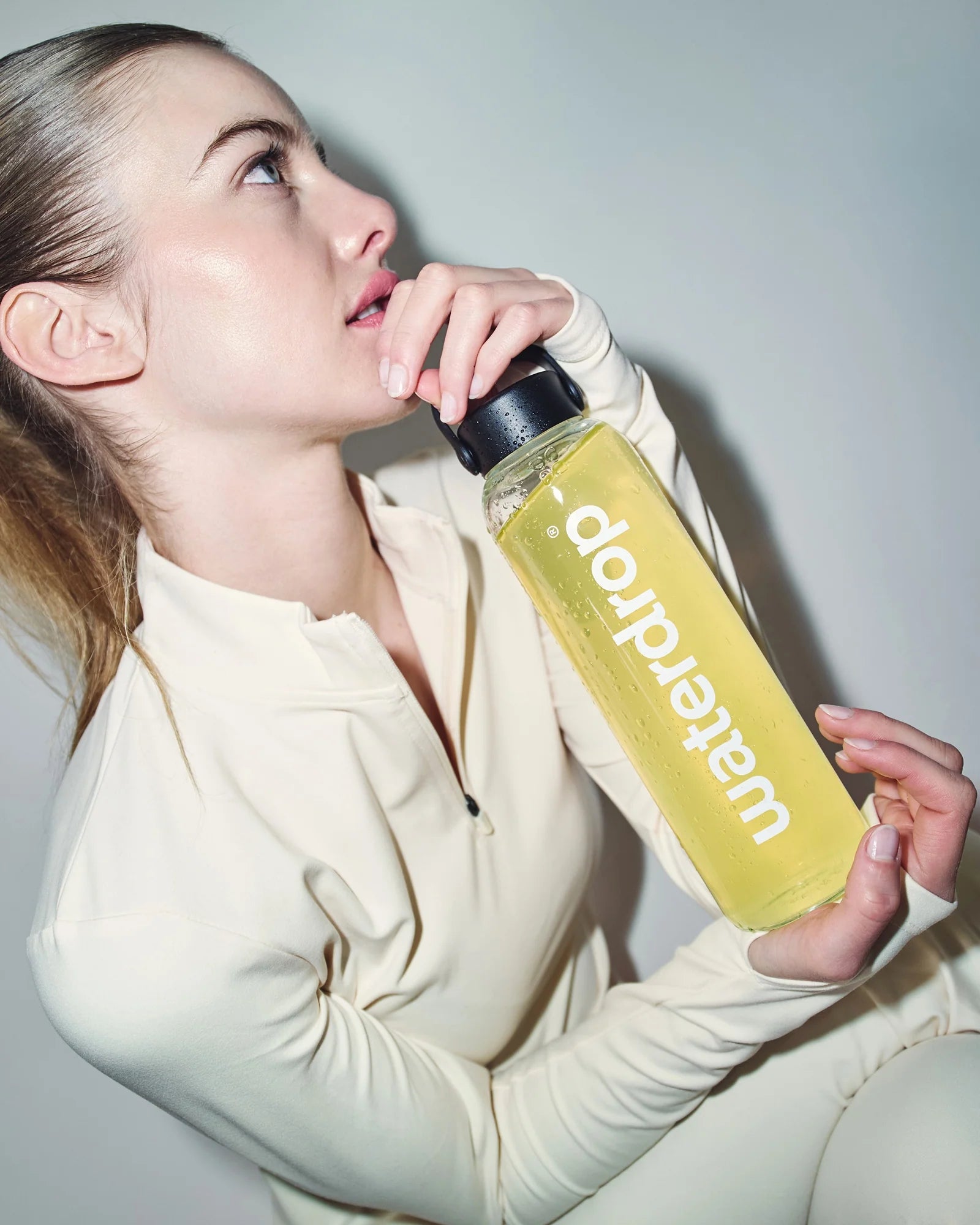 Világos színű sportruhát viselő nő, aki egy sárga Lemon-Mint itallal töltött waterdrop palackot tart, semleges háttér előtt.