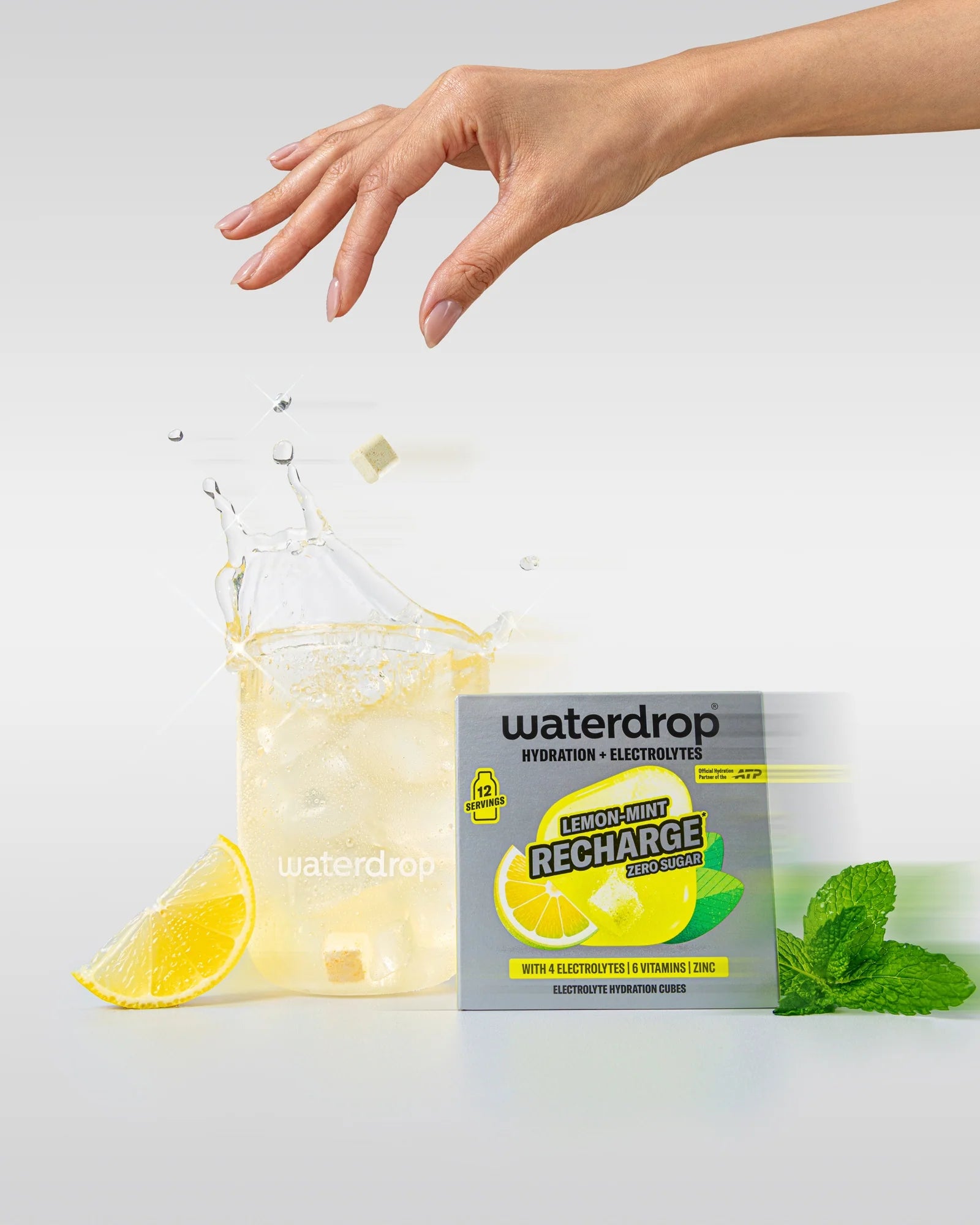 Kéz egy Lemon-Mint italos, jégkockás waterdrop pohár felett, beleeső hidratáló kockával, mellette egy cukormentes Lemon-Mint Recharge doboz 12 adaggal, citromszelet és menta.