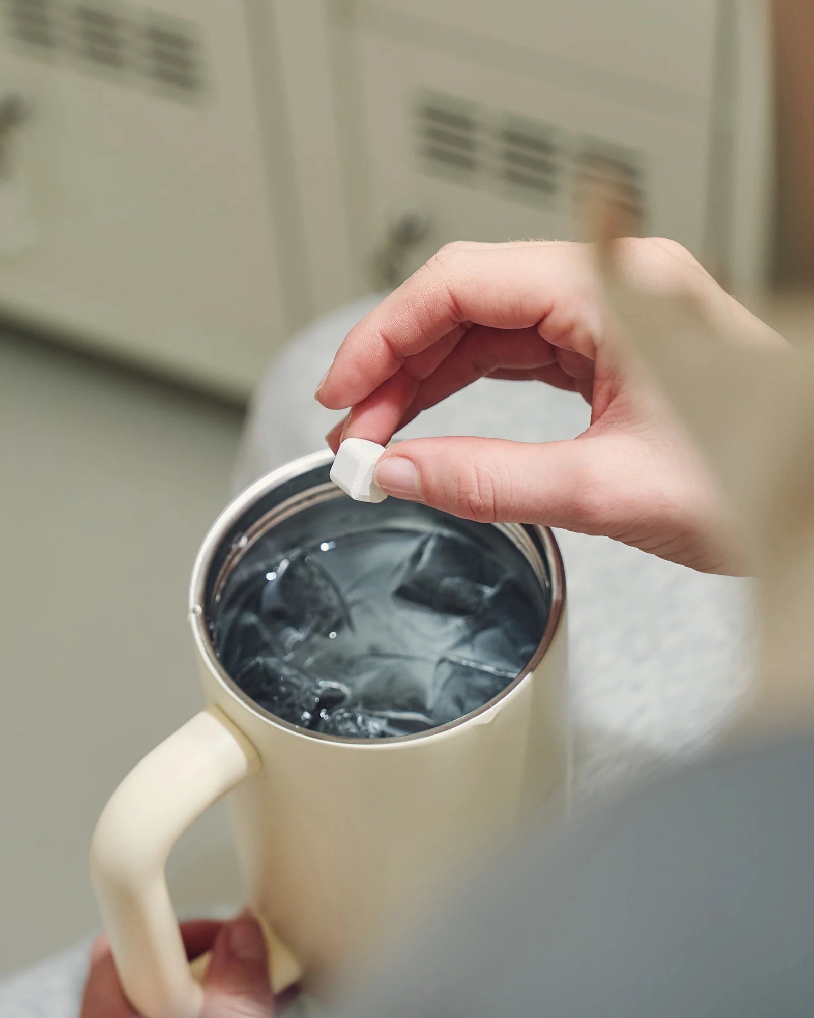 Kéz, amely egy hidratáló kockát tart egy bézs waterdrop termoszbögre fölött, felülnézetből, a bögrében víz és jégkockák láthatók.