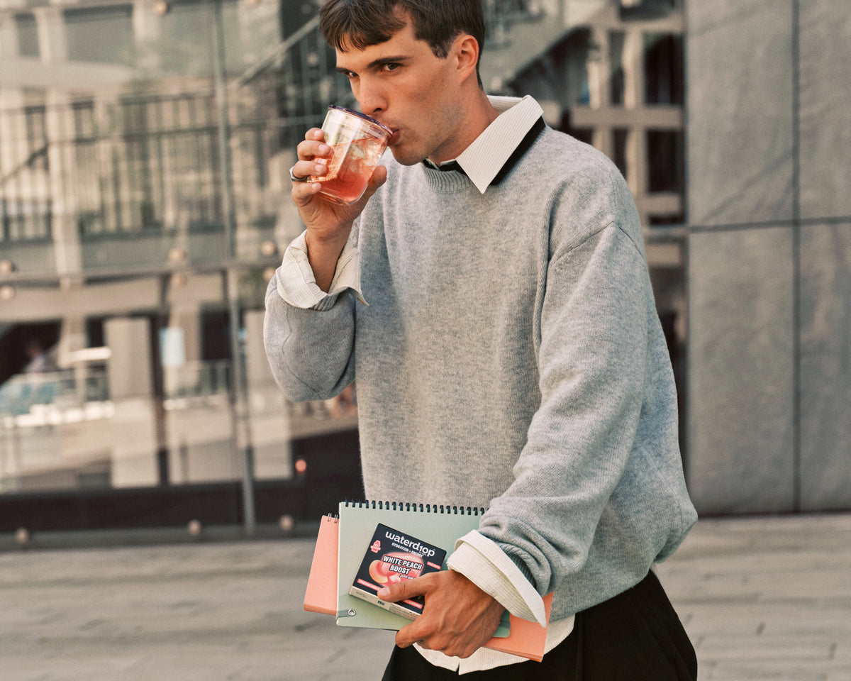 Szürke pulóvert viselő férfi pohárral iszik, miközben kint, városi környezetben noteszt tart.